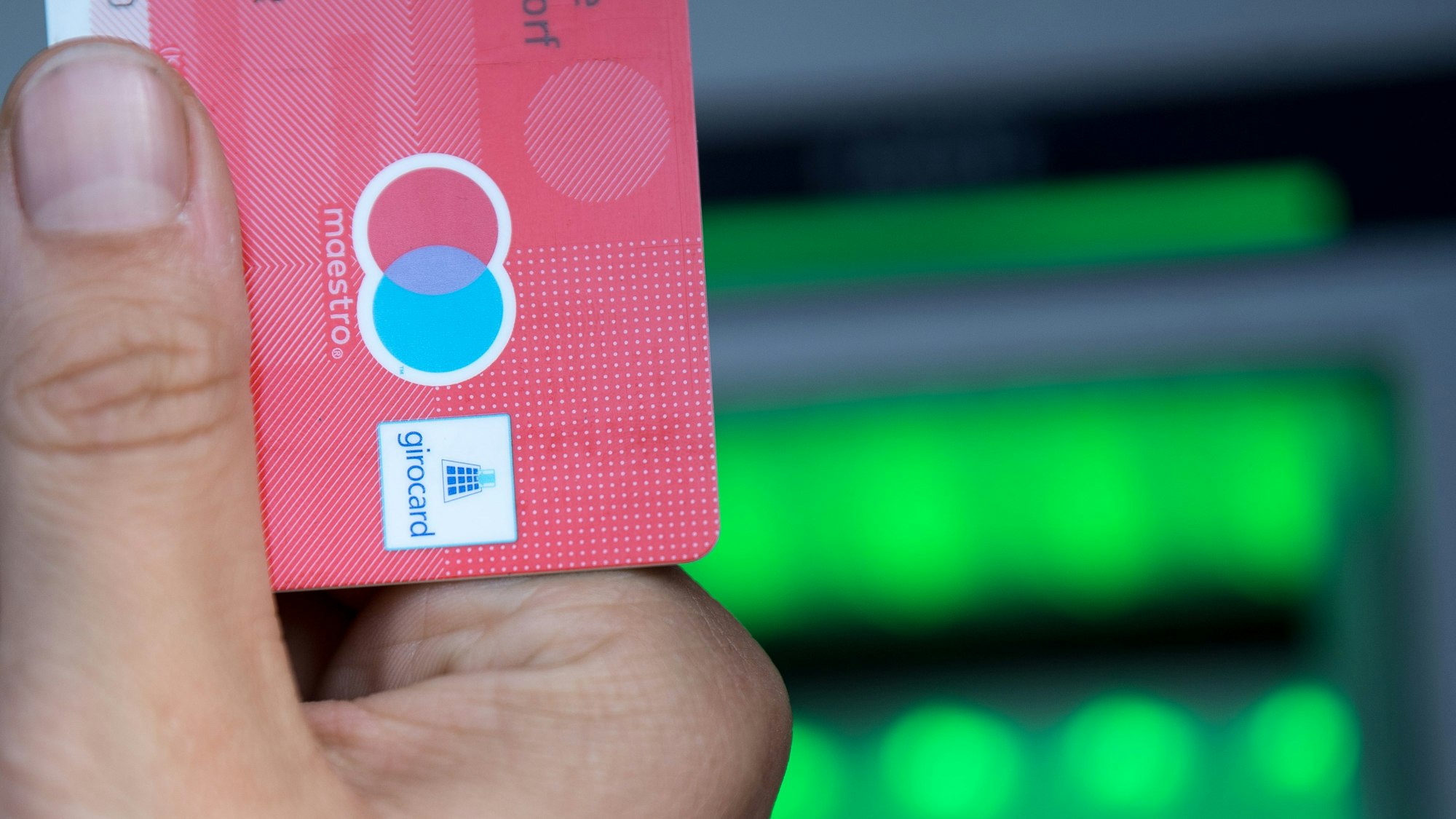 Eine Person hält vor einem Geldautomaten eine Girocard in der Hand. Auf der Karte ist das Maestro-Logo zu sehen.