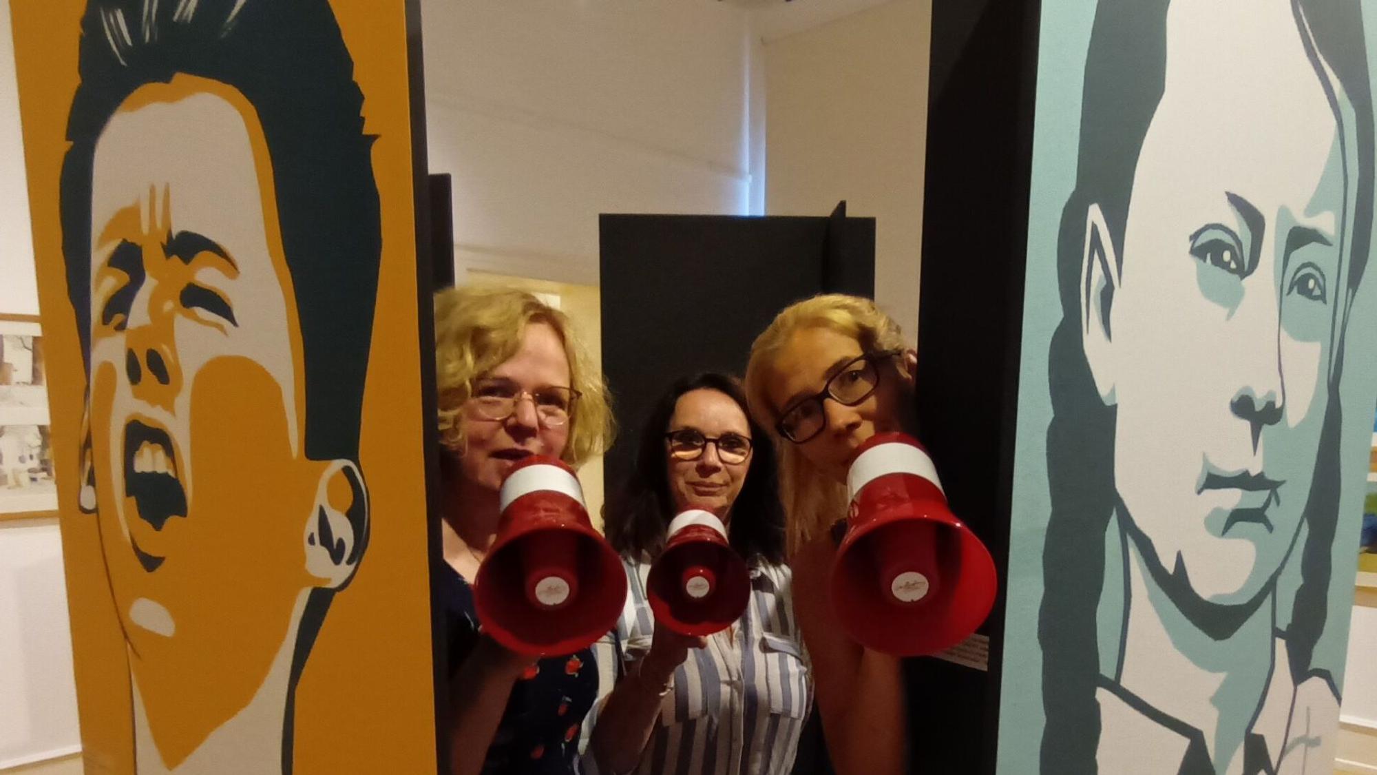 Zwischen zwei Plakaten mit farbigen Kindergesichtern stehen drei Frauen mit Megafon.