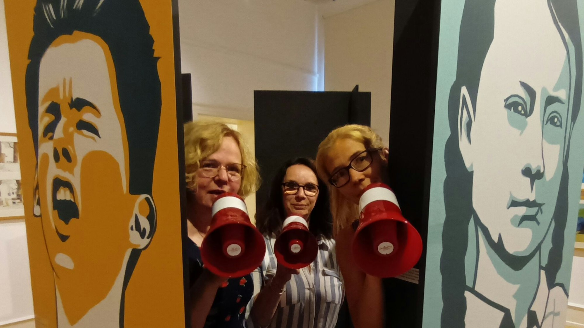 Zwischen zwei Plakaten mit farbigen Kindergesichtern stehen drei Frauen mit Megafon.