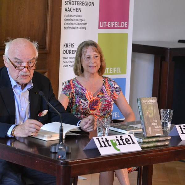 Autor Charles Brauer sitzt mit Gisela Steinhauer an einem Tisch und liest aus seinem Buch.