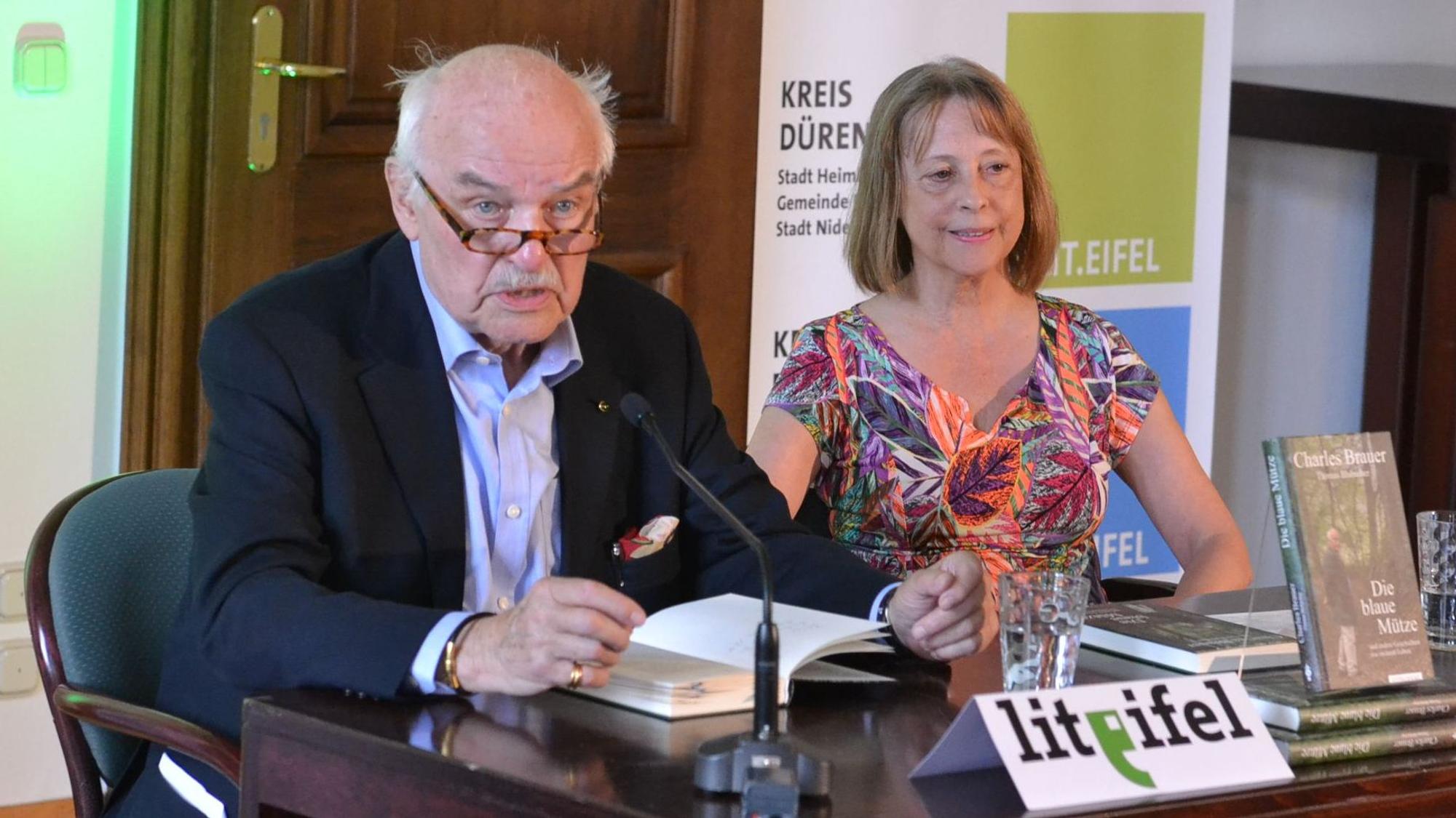 Autor Charles Brauer sitzt mit Gisela Steinhauer an einem Tisch und liest aus seinem Buch.