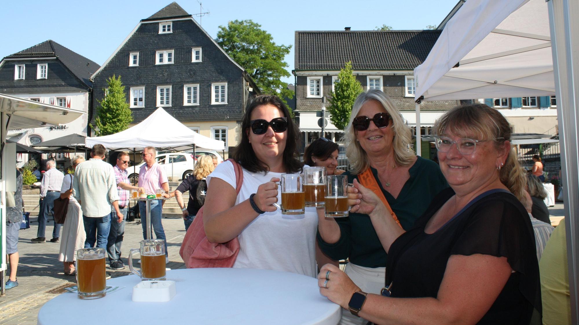 Drei Frauen stehen an einem Stehtisch, lächeln in die Kamera und prosten sich mit einem Bier zu.