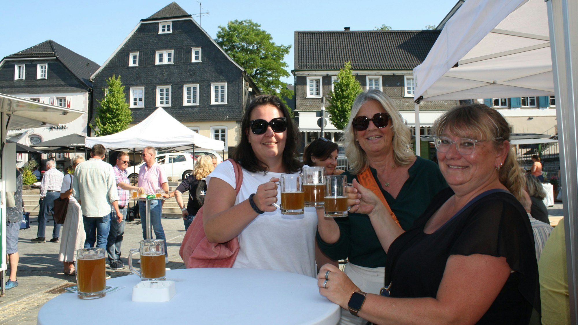 Drei Frauen stehen an einem Stehtisch, lächeln in die Kamera und prosten sich mit einem Bier zu.