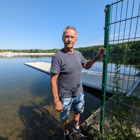 Kunze steht in kurzer Hose und T-Shirt neben dem Zaun. Im Hintergrund ist der Bootssteg zu sehen.