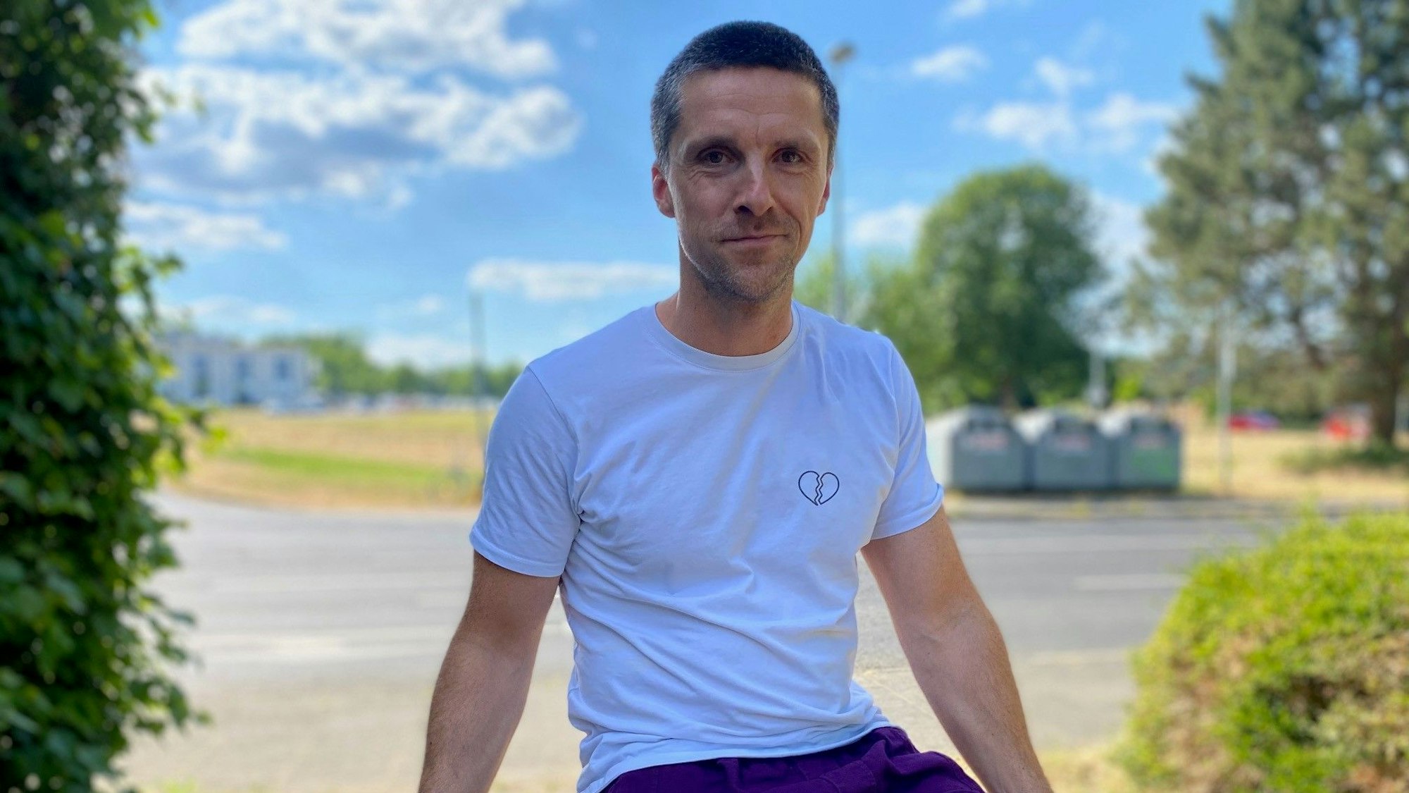 Bela trägt ein weißes T-Shirt, eine lilane Hose und lächelt in die Kamera.