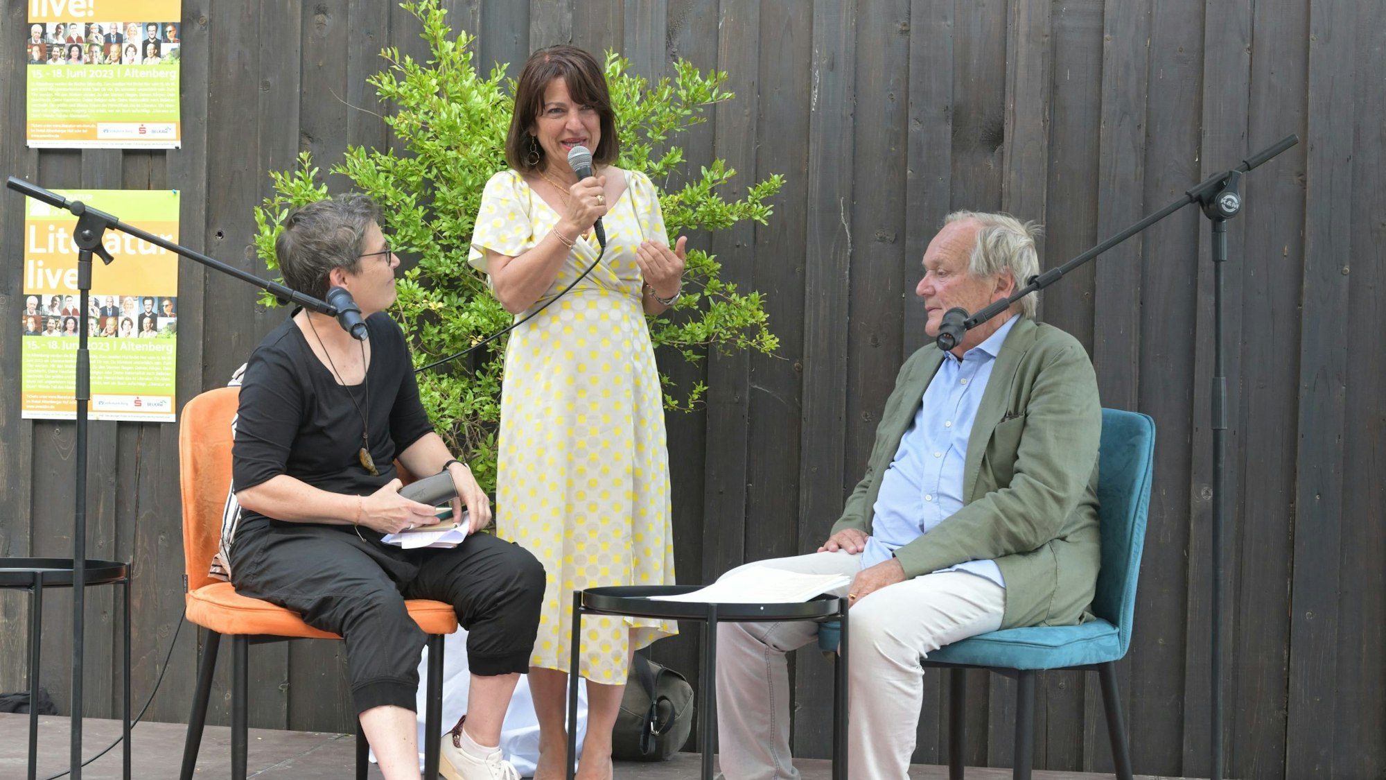 Eine Frau und ein Mann sitzen auf Stühlen vor Mikros, eine weitere Frau steht dazwischen und spricht in ein Mikrofon.
