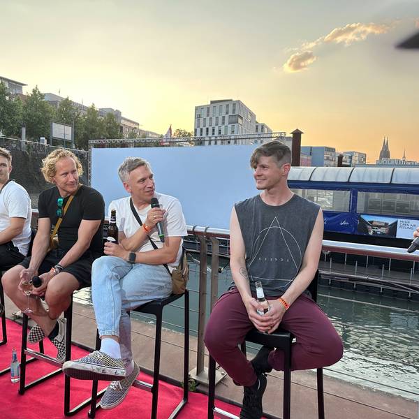 Die Band sitzt auf Hockern, Basti Campmann spricht ins Mikro. Im Hintergrund der Rheinauhafen.