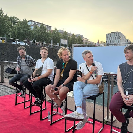 Die Band sitzt auf Hockern, Basti Campmann spricht ins Mikro. Im Hintergrund der Rheinauhafen.
