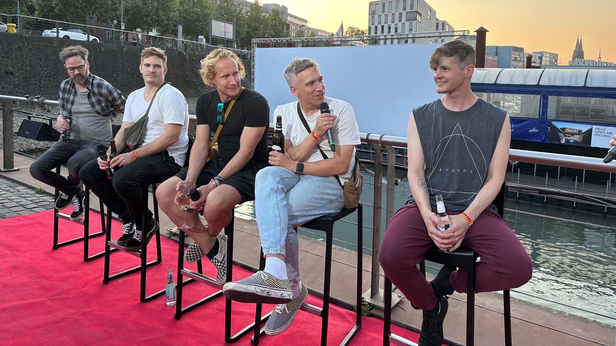 Die Band sitzt auf Hockern, Basti Campmann spricht ins Mikro. Im Hintergrund der Rheinauhafen.