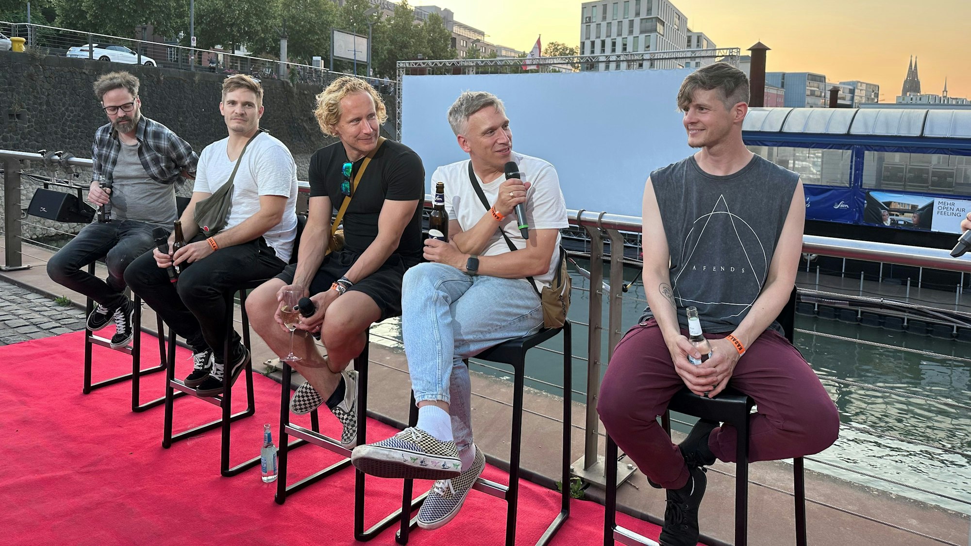 Die Band sitzt auf Hockern, Basti Campmann spricht ins Mikro. Im Hintergrund der Rheinauhafen.