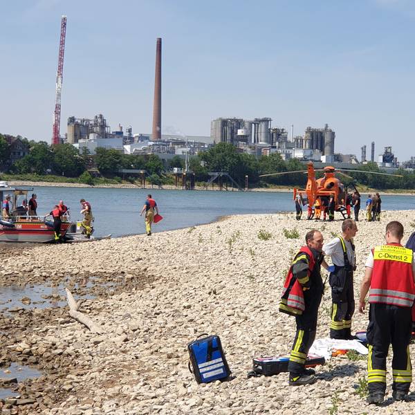 Rettungskräfte stehen am Strand des Rheins in Niederkassel. Vor dem Ufer liegt ein Boot, ein Rettungshubschrauber steht weiter hinten.