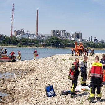Rettungskräfte stehen am Strand des Rheins in Niederkassel. Vor dem Ufer liegt ein Boot, ein Rettungshubschrauber steht weiter hinten.