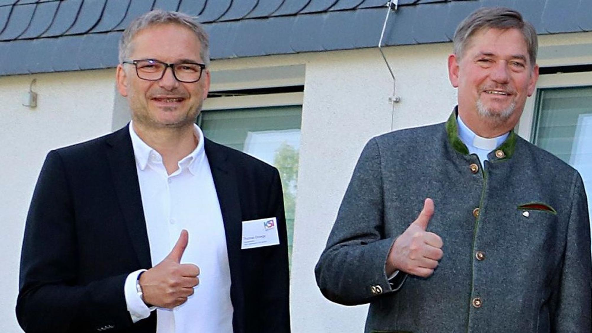 Zwei Männer stehen mit nach oben gestrecktem Daumen vor einem Gebäude.