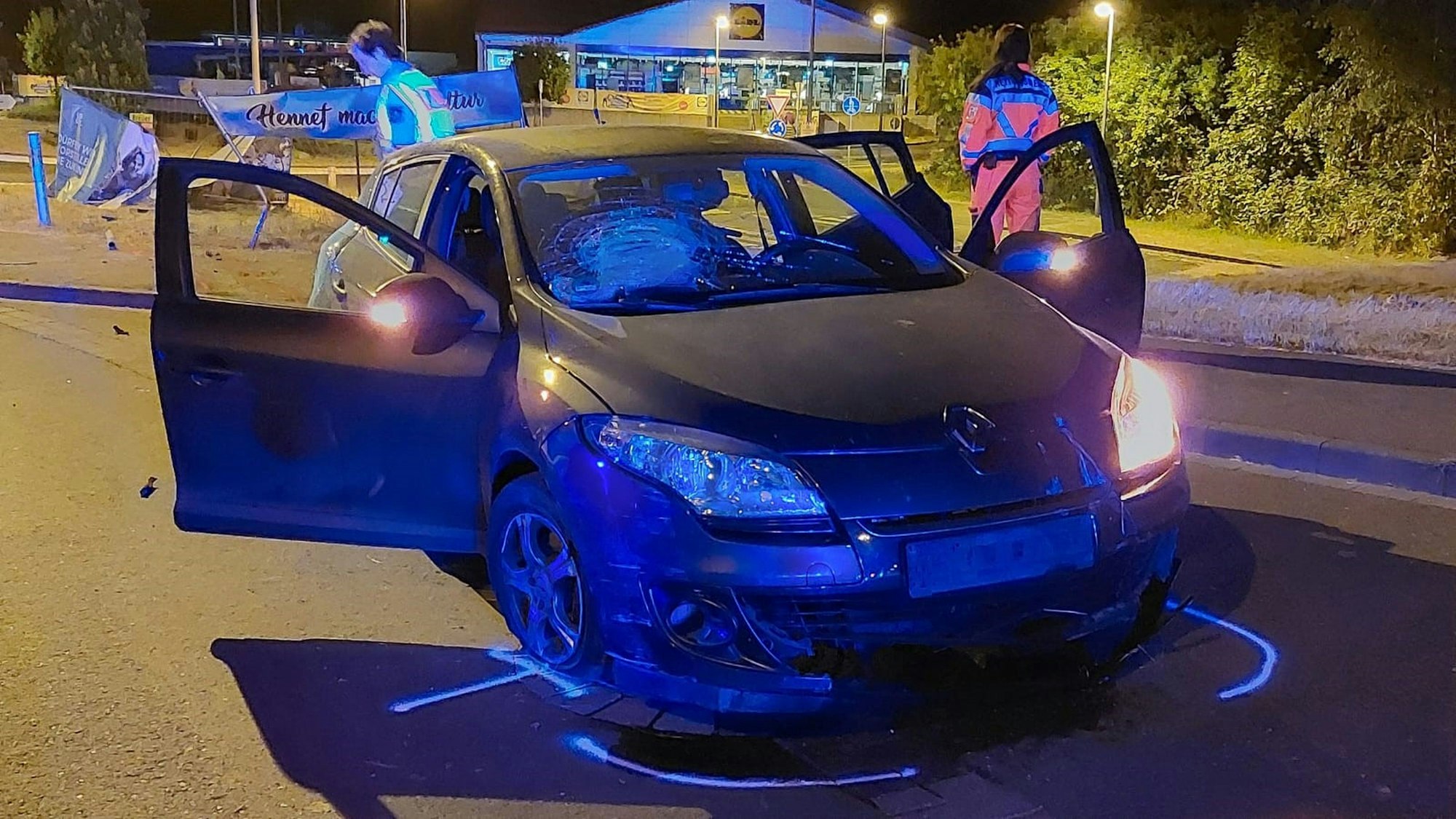 Ein Pkw mit einer zersplitterten Frontscheibe steht auf einer nächtlichen Straße, um ihn sind Markierungen aufgesprüht. Im Hintergrund steht ein zerstörter Bauzaun.