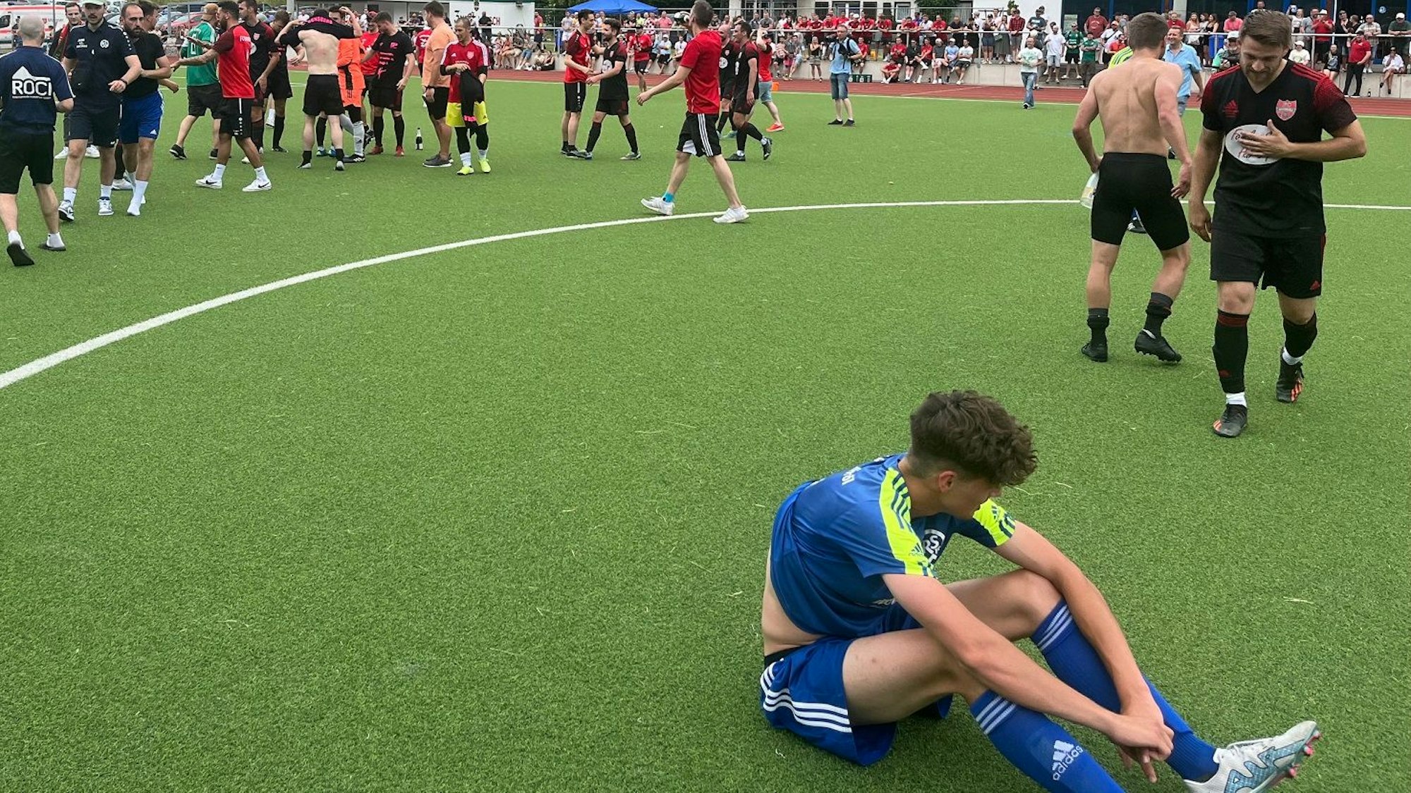 Zu sehen ist ein enttäuschter Fußballer, der auf dem Rasen sitzt, im Hintergrund feiert die gegnerische Mannschaft.