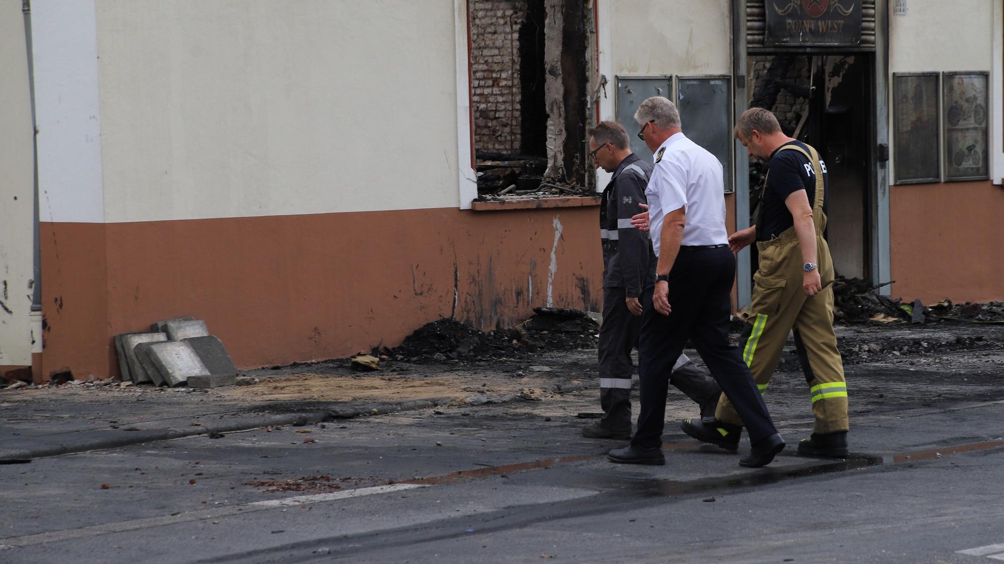 Drei Männer in Funktionskleidung gehen mit gesenkten Blicken auf das ausgebrannte Haus in Sankt Augustin zu.