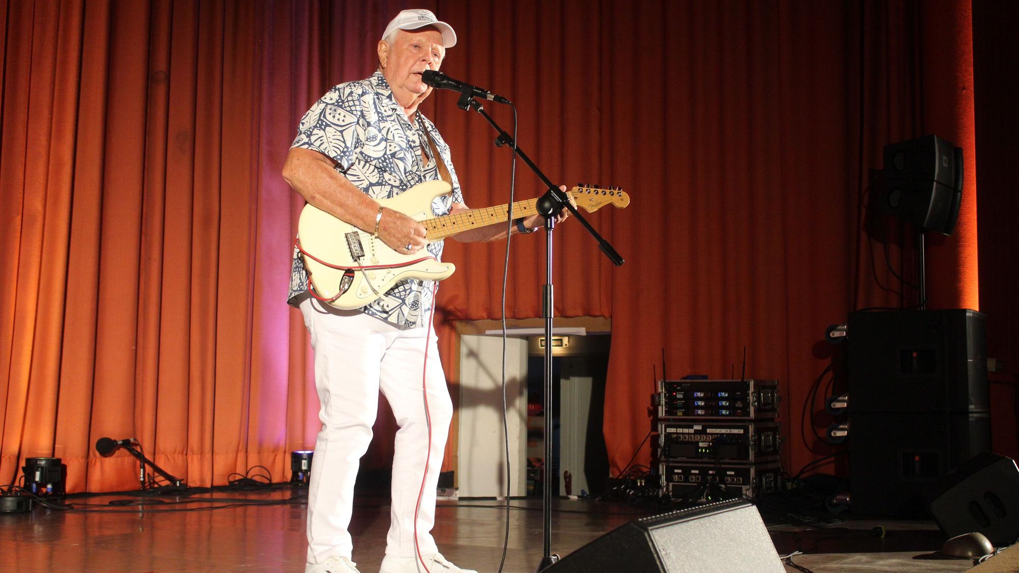Schlagerstar Graham Bonney steht mit Gitarre auf einer Bühne und singt in ein Standmikro.