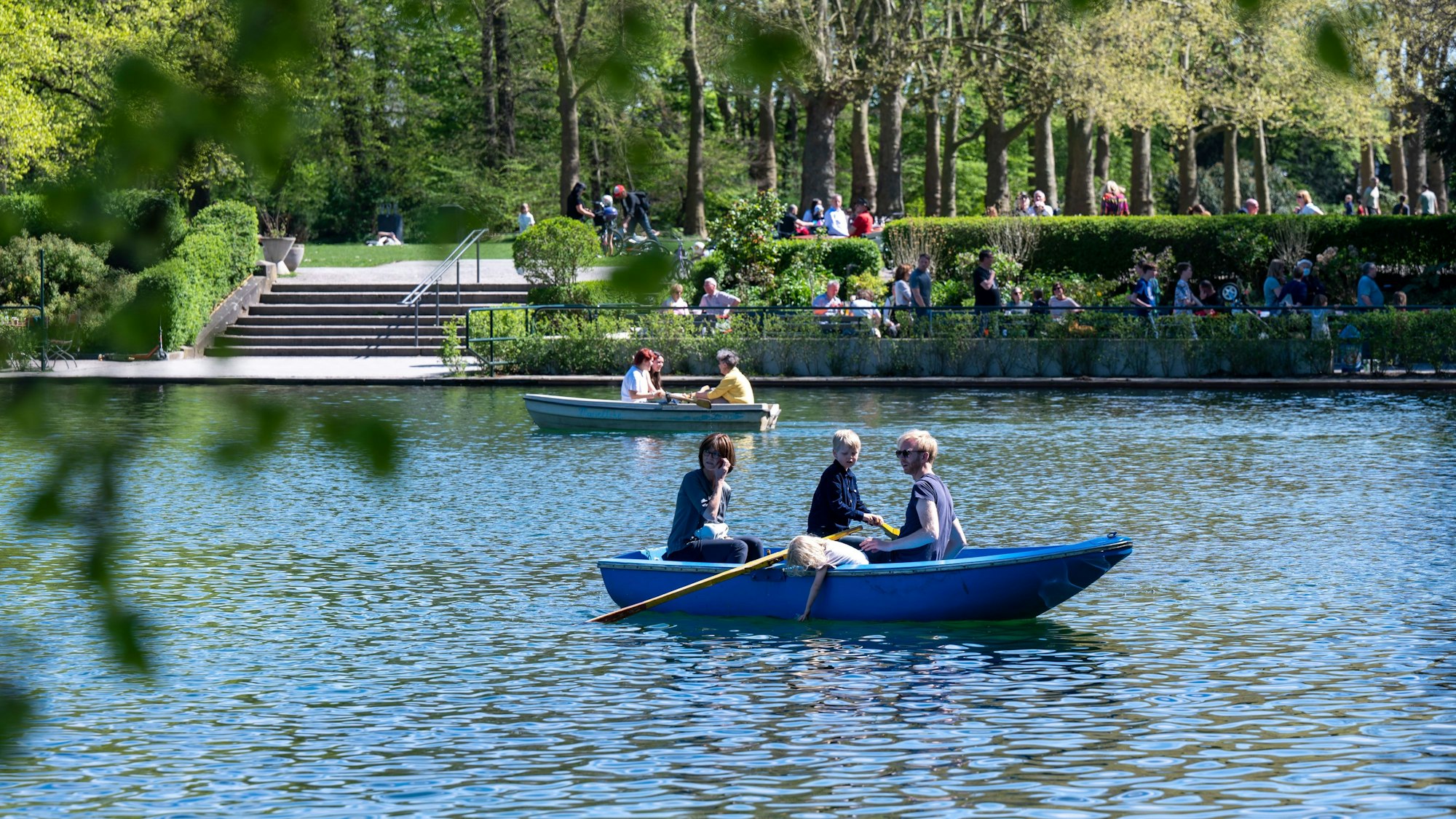 zwei Ruderboote fahren auf einem Weiher, im Hintergrund sitzen Menschen auf einem Weg am Ufer