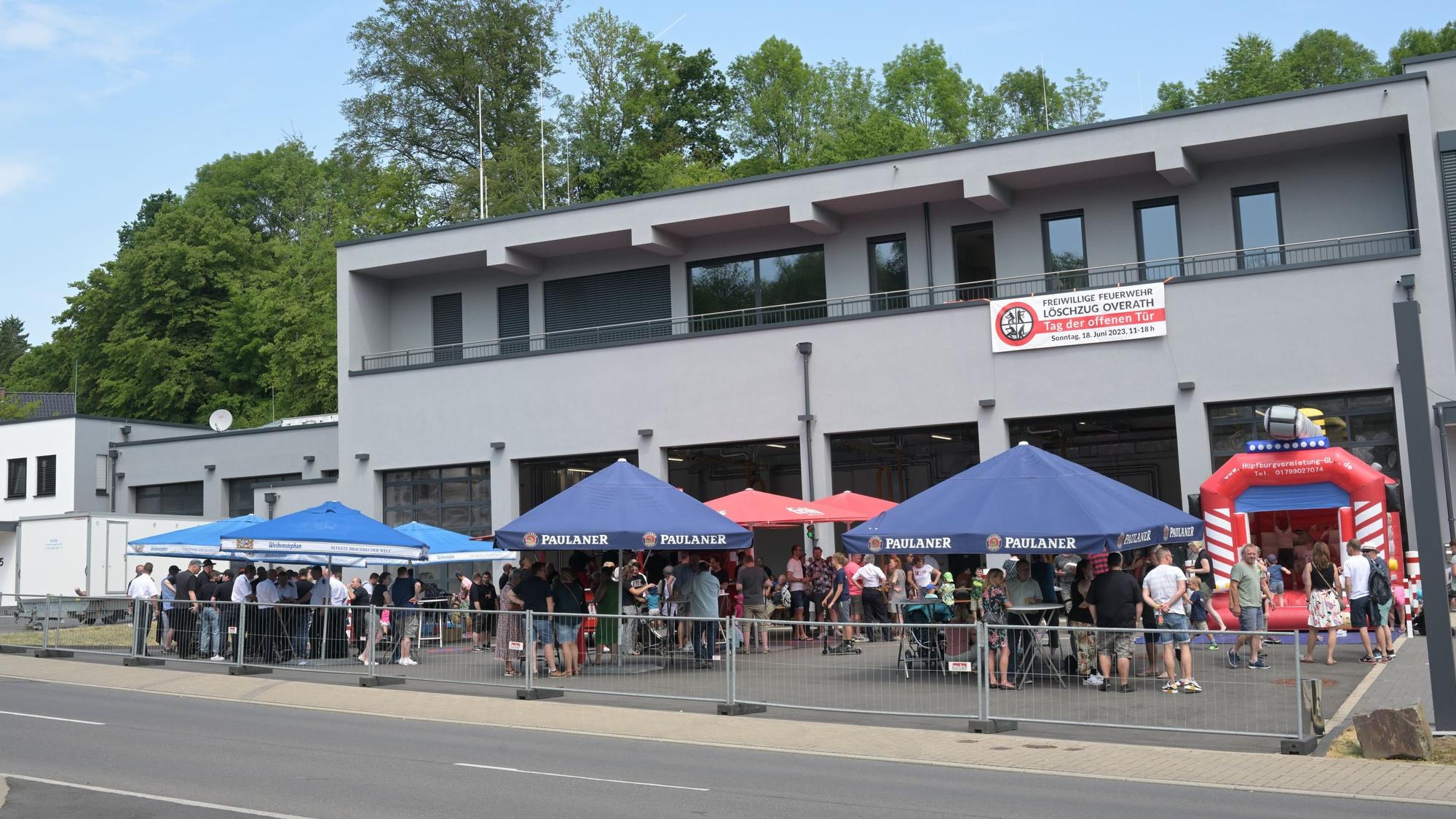 Schirme stehen vor dem Feuerwehrhaus Overath, darunter stehen Menschen zusammen, daneben ist eine Hüpfburg zu sehen.