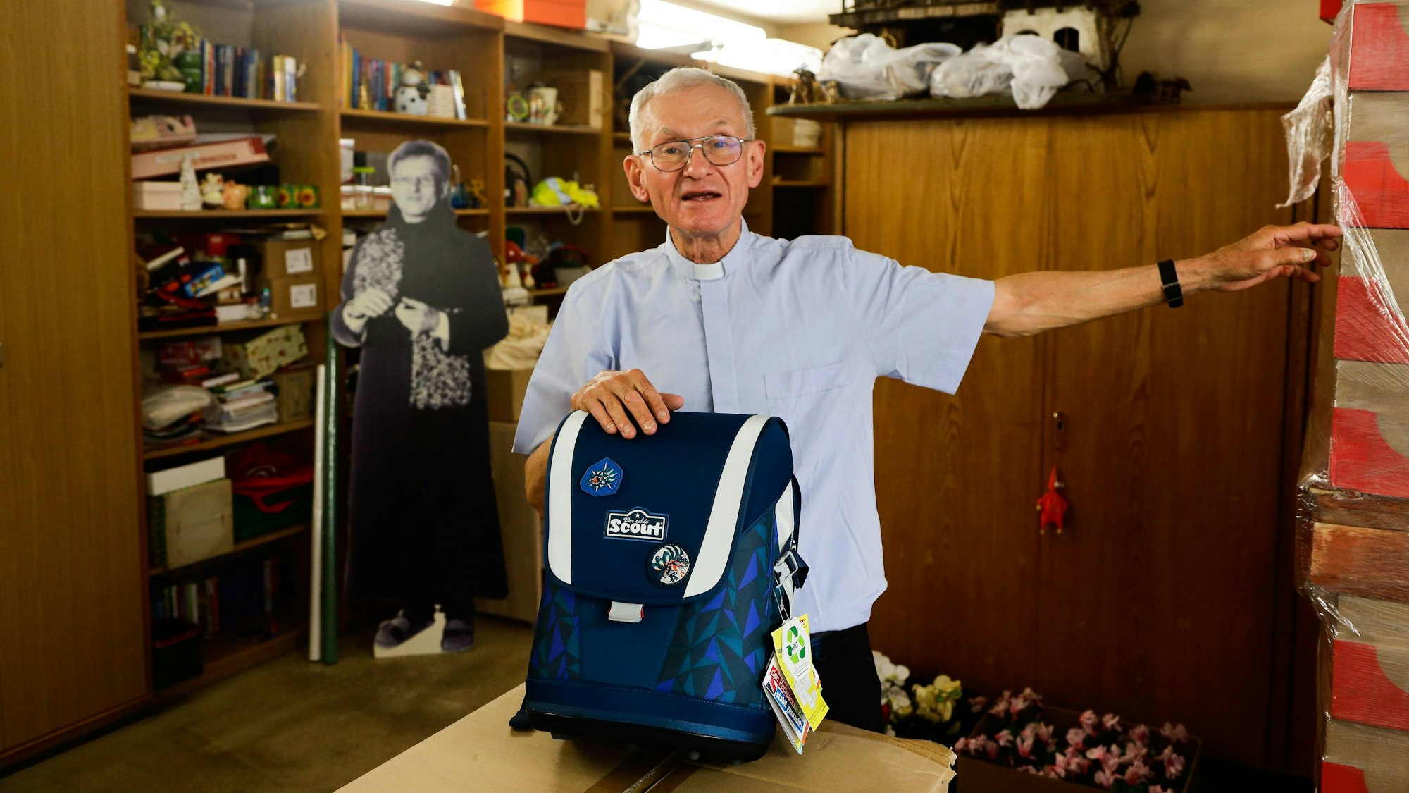 Pfarrer Franz Meurer steht in einem Kellerraum und präsentiert einen Schulranzen