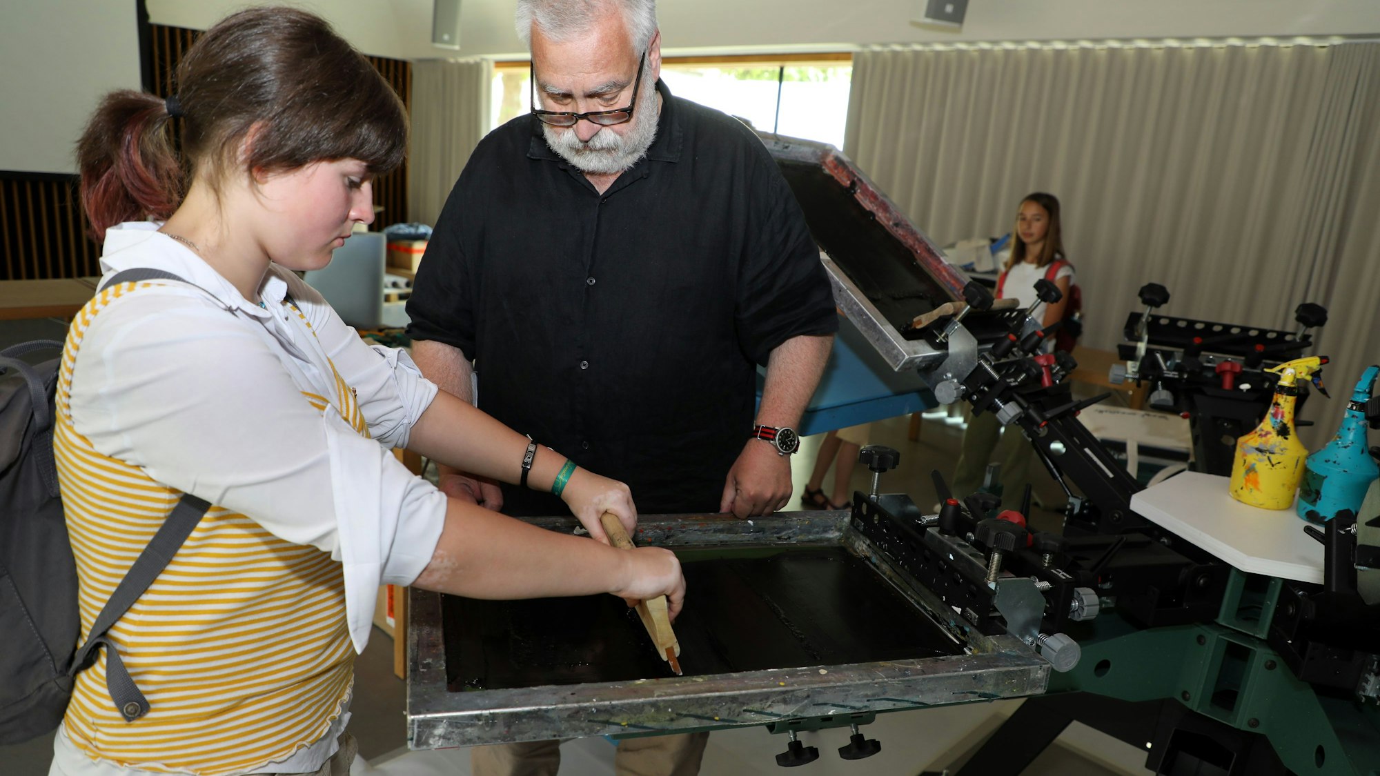 Ein Mädchen und ein Mann stehen an einer Siebdruckplatte und Streichen Farbe mit einem Rakel über das Sieb.