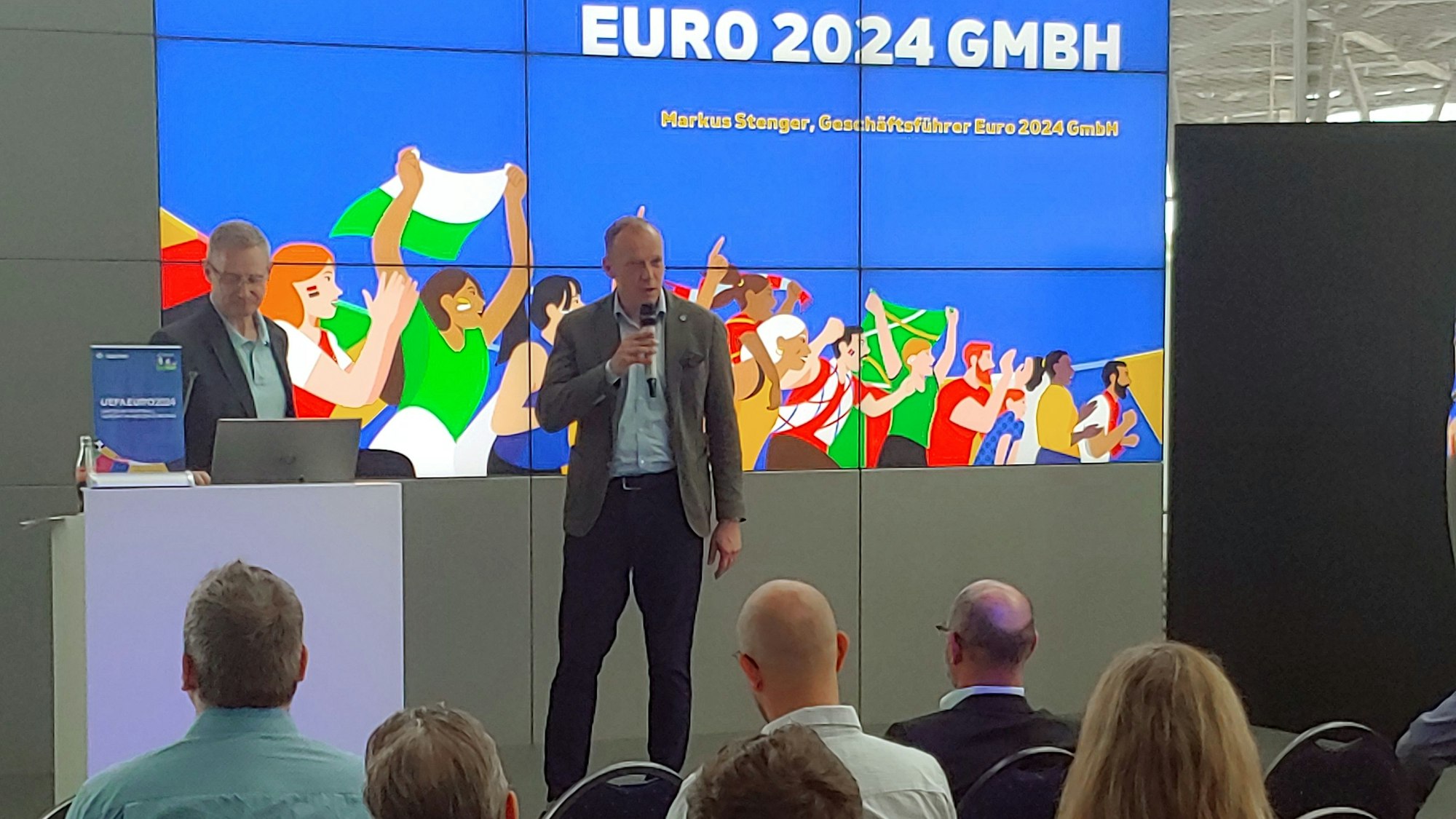 Zwei Männer auf der Bühne, davor Publikum auf Stühlen