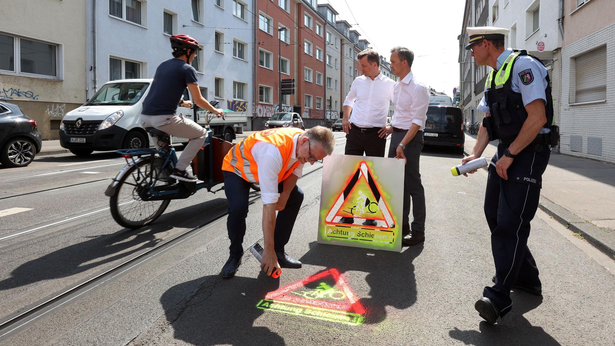 Ein KVB-Mitarbeiter besprüht die Straße mit einem Warnsymbol. Im Hintergrund halten Polizeipräsident Falk Schnabel und Verkehrsdezernent Ascan Egerer das Piktogramm hoch.