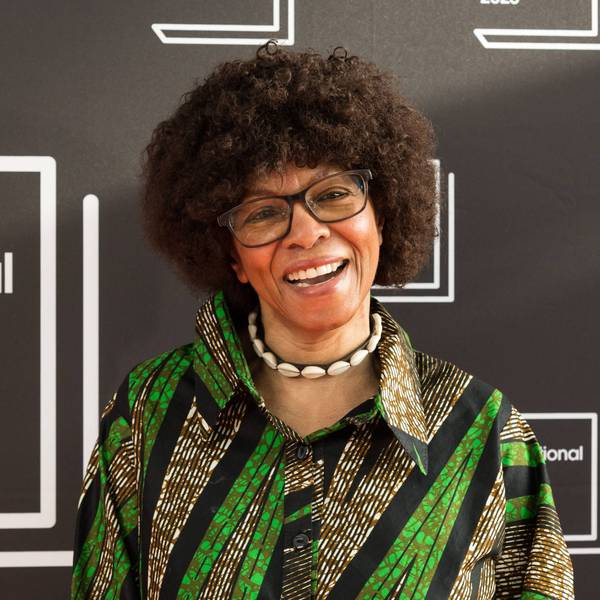 Margaret Busby trägt eine Brille, eine Muschelkette und ein grün gemustertes Hemd. Sie lacht in die Kamera.