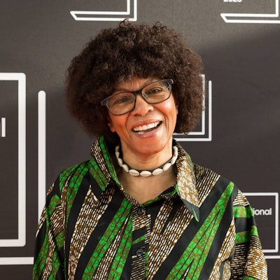 Margaret Busby trägt eine Brille, eine Muschelkette und ein grün gemustertes Hemd. Sie lacht in die Kamera.