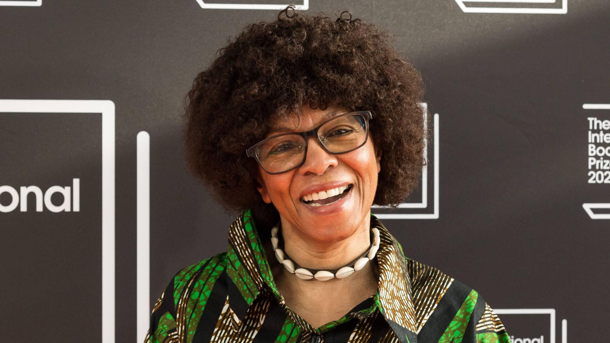 Margaret Busby trägt eine Brille, eine Muschelkette und ein grün gemustertes Hemd. Sie lacht in die Kamera.