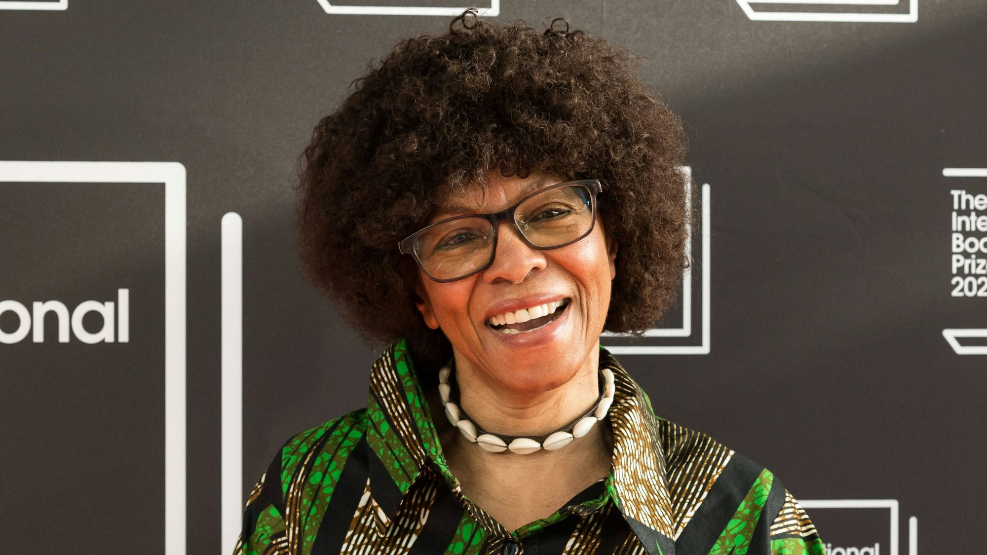 Margaret Busby trägt eine Brille, eine Muschelkette und ein grün gemustertes Hemd. Sie lacht in die Kamera.