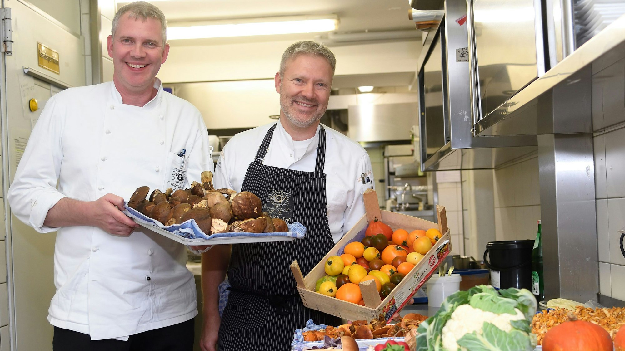 Alejandro und Christopher Wilbrand stehen mit Obst, Gemüse und Pilzen in der Küche ihres Restaurants.