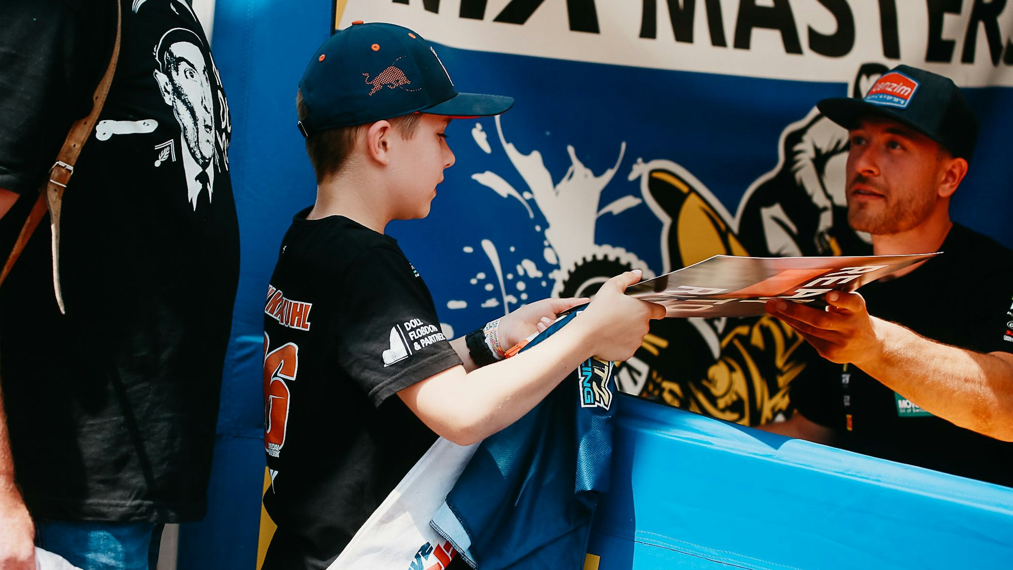 Ein Motocross-Fahrer überreicht einem kleinen Jungen ein Poster mit seiner Unterschrift.