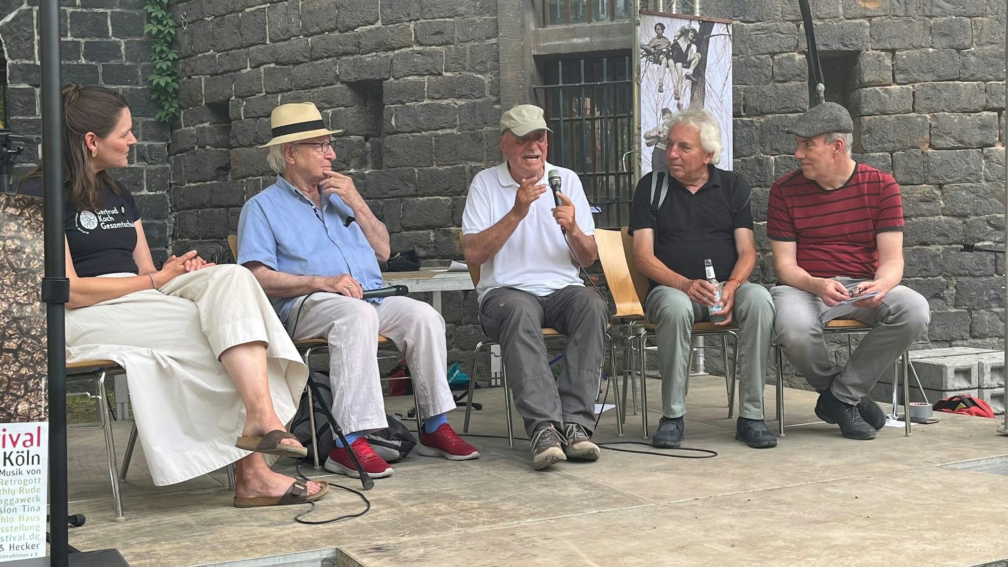 Eine Frau und vier Männer sitzen auf Stühlen nebeneinander, einer hält ein Mikrofon und gestikuliert.