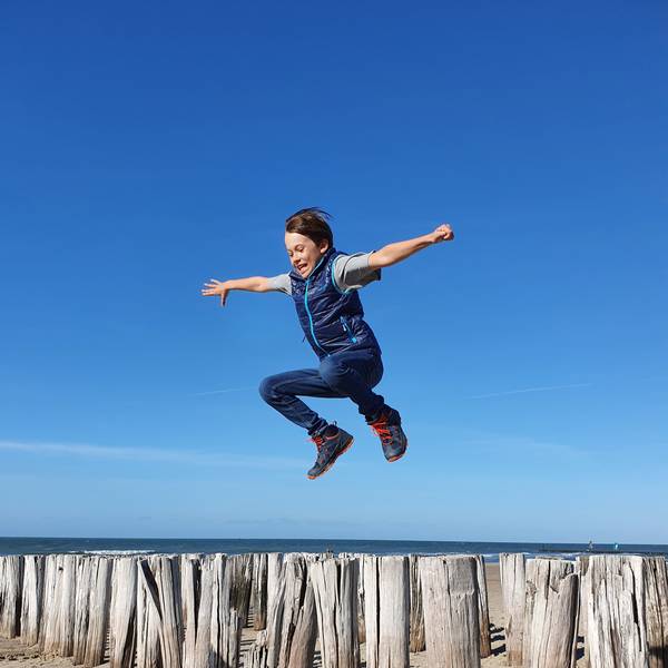 Ein Junge springt über Hölzer an einem Strand vor blauem Himmel und Meereshorizont.
