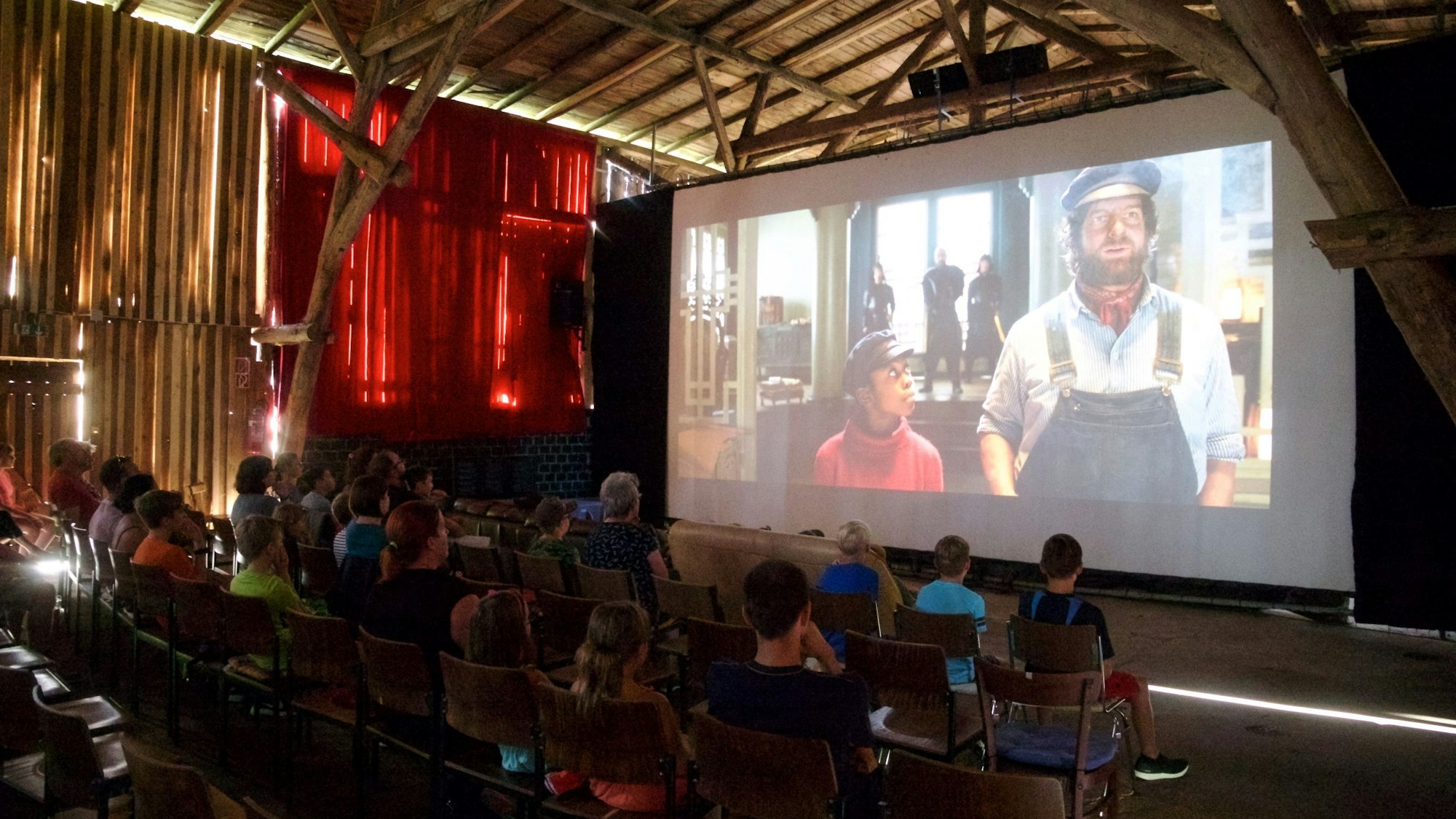 Das Bild zeigt eine große Leinwand, auf der ein Film gezeigt wird. Davor sitzt das Publikum auf Stühlen.