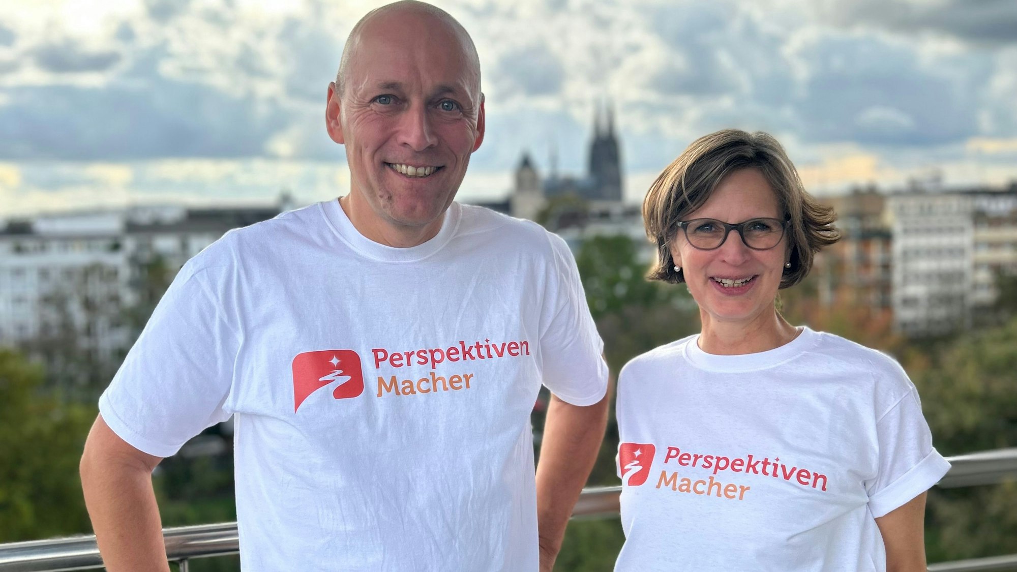 Zwei Menschen im weißen T-Shirt vor Kölner Kulisse.
