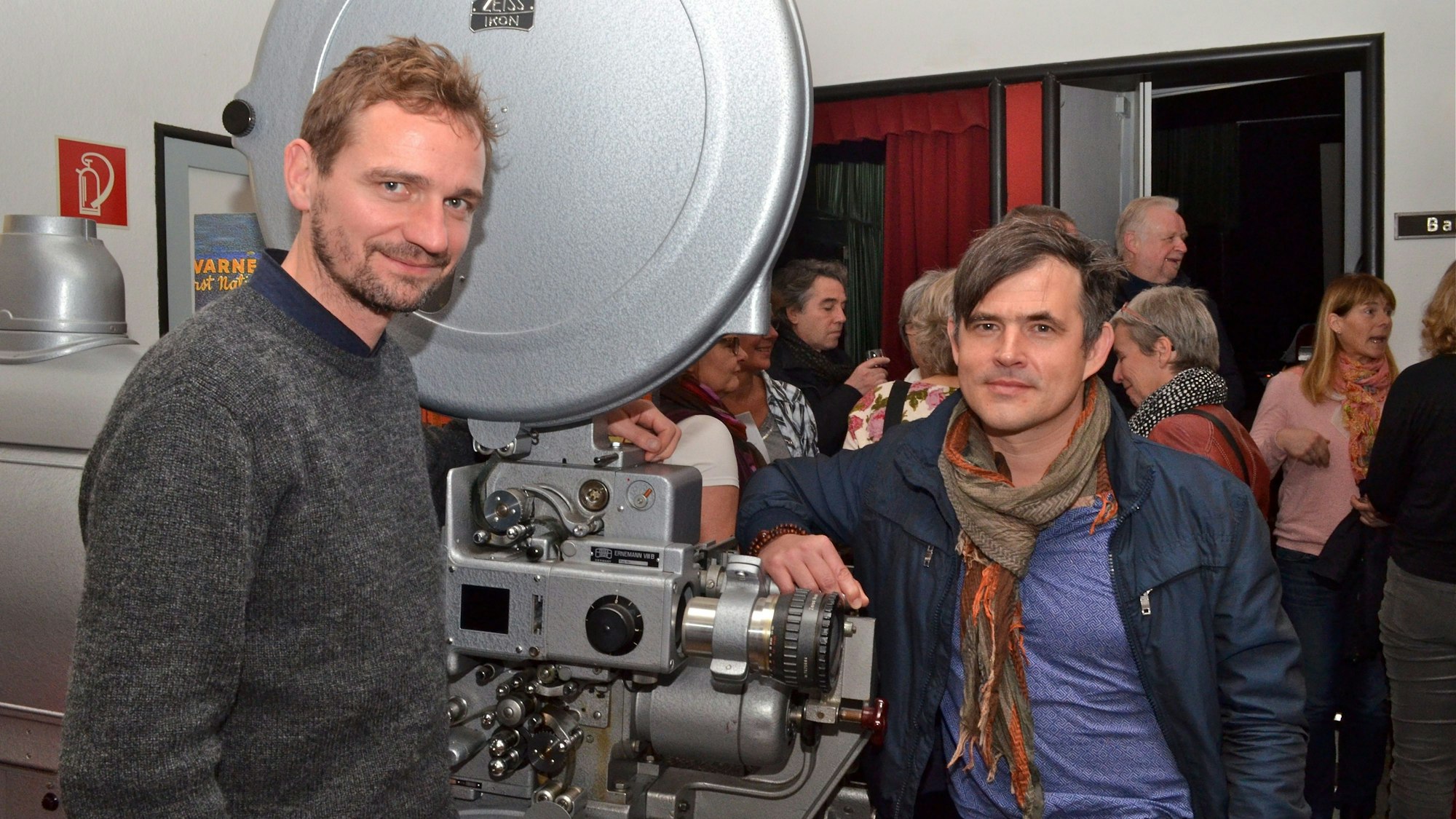 Schauspieler Maik Solbach (l.) steht mit Regisseur Oliver Haffner vor einem Kino-Projektor beim Filmfestspielchen Hennef in 2014. Im Hintergrund sind Menschen zu sehen.