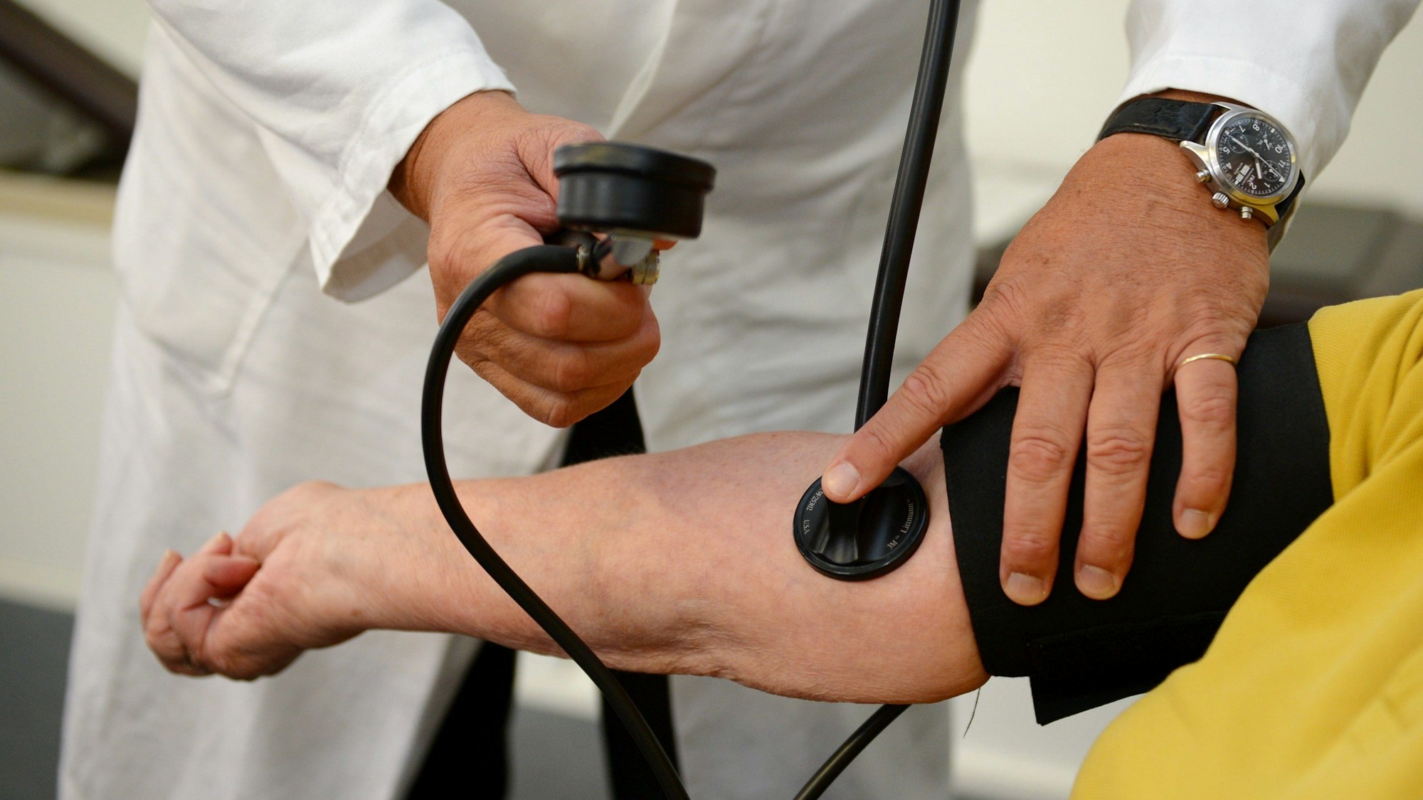 Ein Arzt misst den Blutdruck einer Patientin oder eines Patienten. Von den Personen sind lediglich der Arm des Patienten und die Hände des Arztes zu sehen.