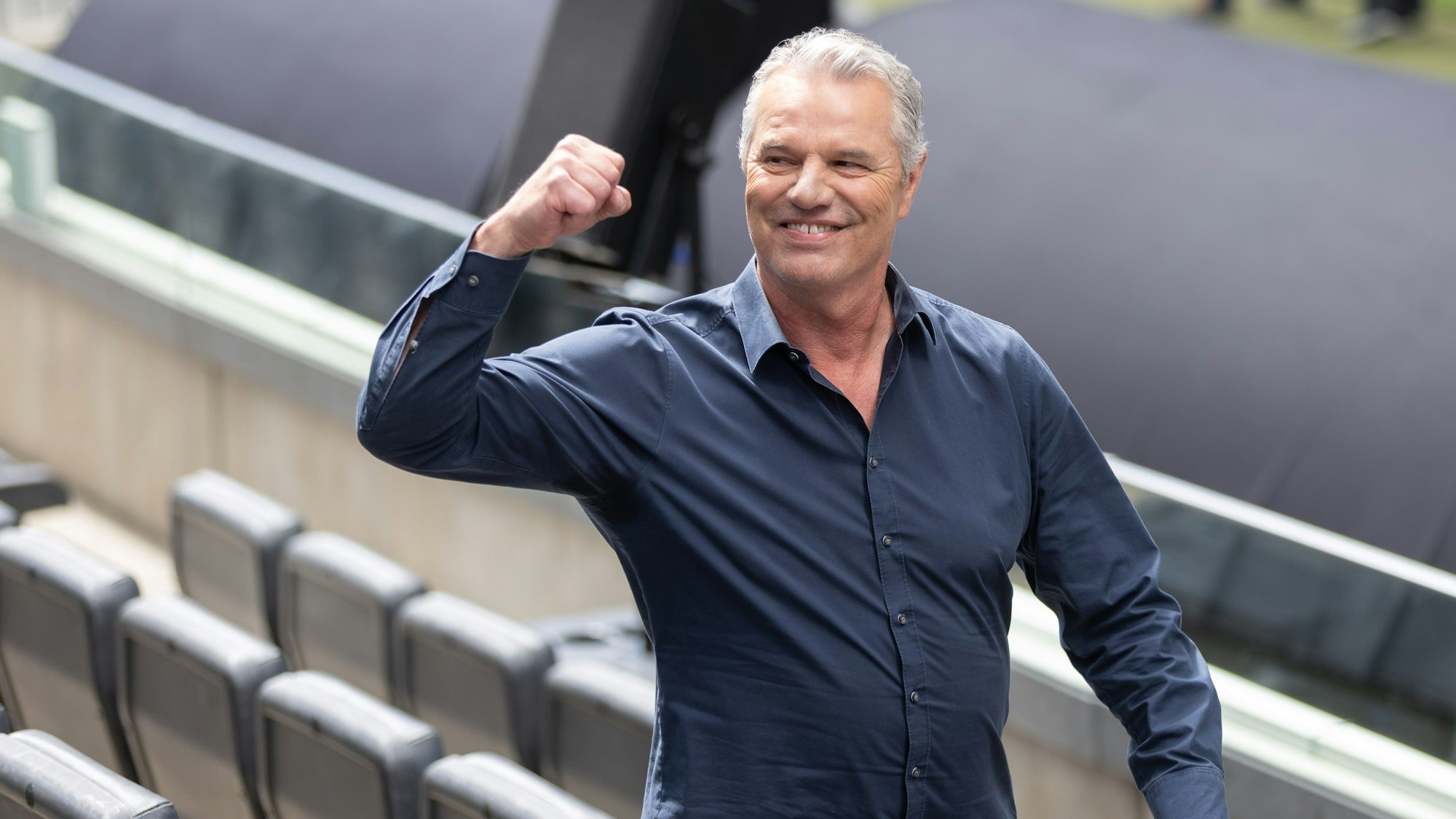 Fernsehmoderator Jan Stecker steht auf der Zuschauertribüne eines Stadions und reckt eine Faust. Er schaut dabei an der Kamera vorbei.