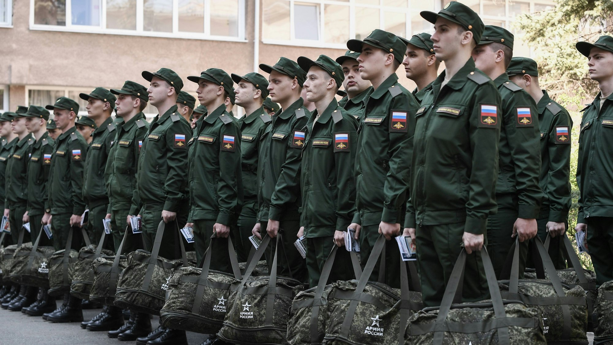 Wehrpflichtige stehen in Formation, bevor sie die Stadt verlassen, um in der russischen Armee zu dienen. Sie tragen grüne Uniform und haben eine Tasche in der Hand.