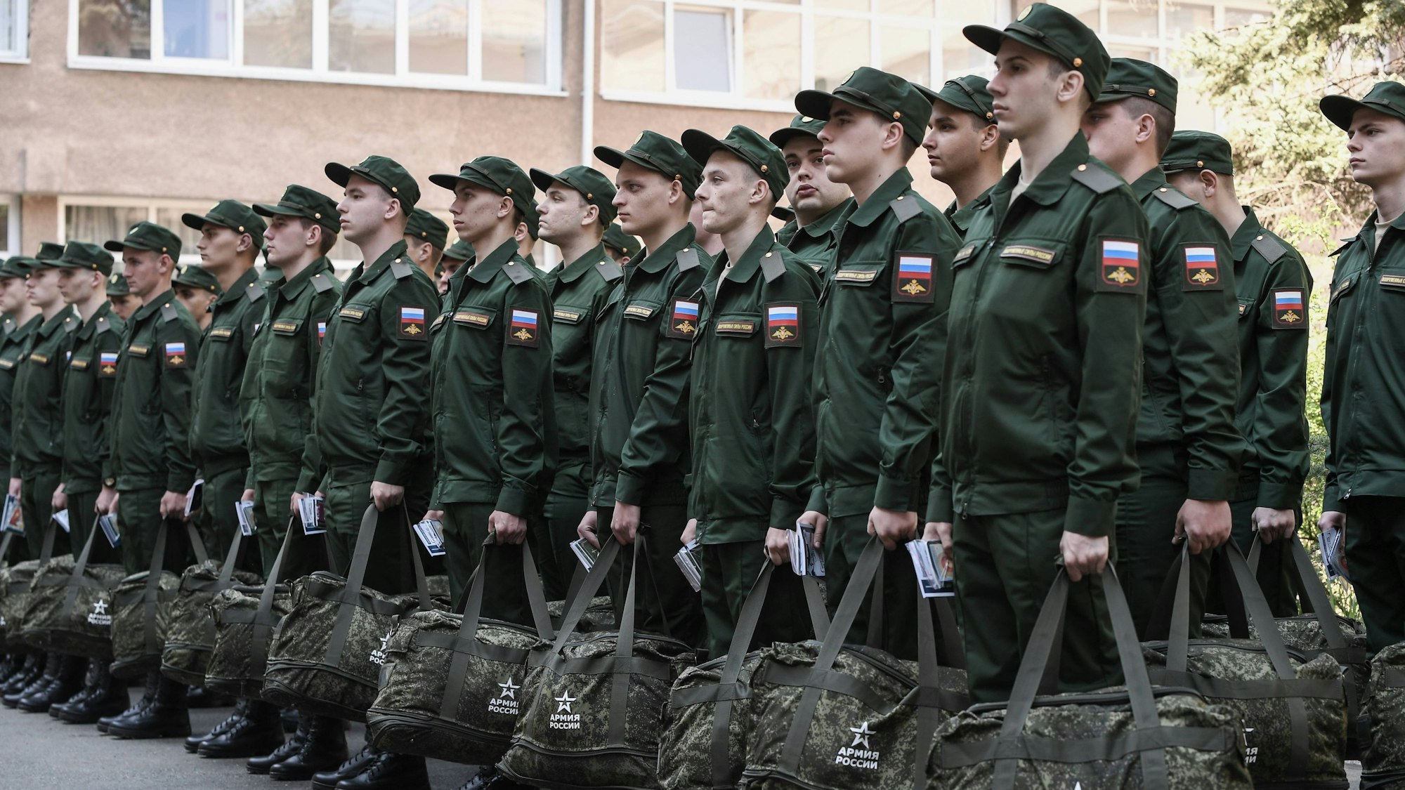 Wehrpflichtige stehen in Formation, bevor sie die Stadt verlassen, um in der russischen Armee zu dienen. Sie tragen grüne Uniform und haben eine Tasche in der Hand.