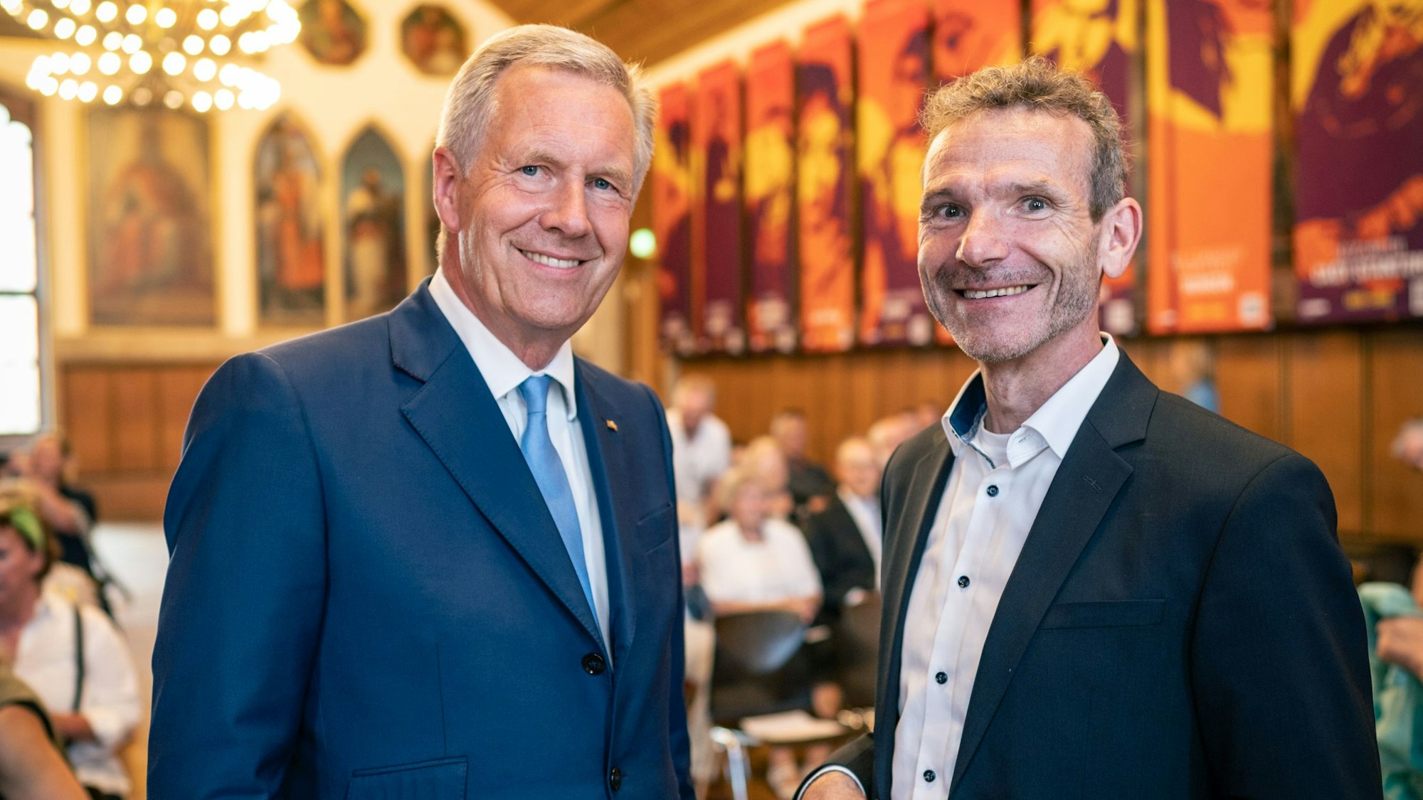 Christian Wulff trägt einen blauen Anzug mit Krawatte, Joachim Frank trägt einen schwarzen Anzug. Beide lachen in die Kamera.