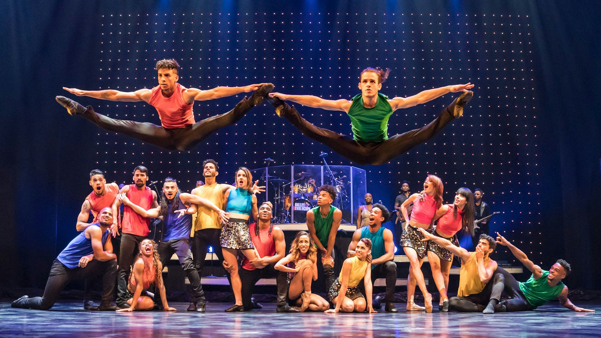 Zwei Tänzer springen in die Höhe und machen einen Spagat, im Hintergrund viele weitere, die sitzend oder stehen ihre Kollegen bewundern. Sie tragen bunte Shirts.