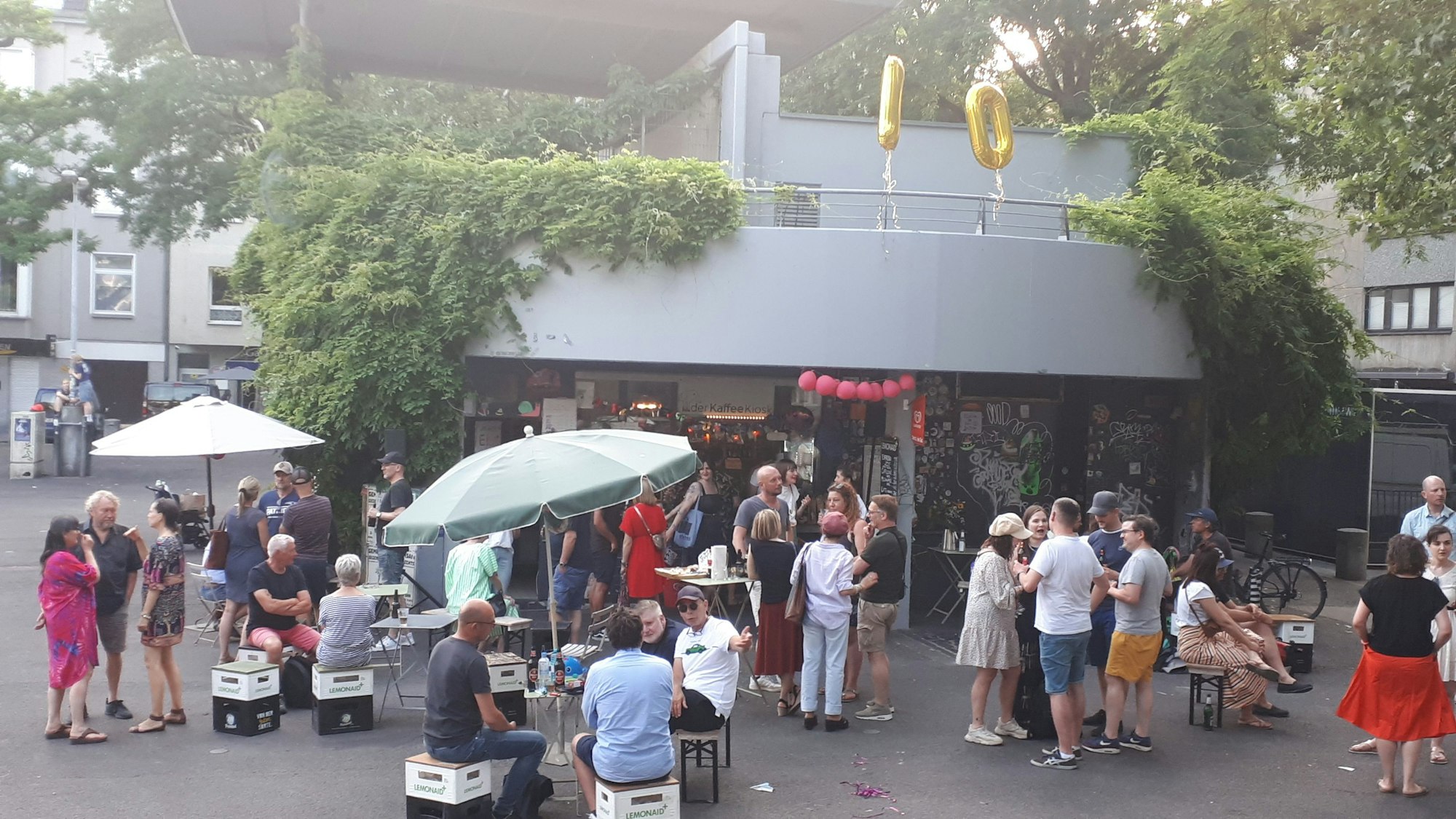 Viele Menschen stehen oder sitzen auf Hockern auf einem Platz vor einem Kiosk, der in einem Betongebäude untergebracht ist.
