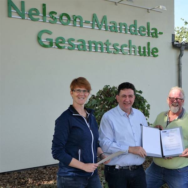 Vier Personen halten einen Vertrag vor dem Gebäude der Nelson-Mandela-Gesamtschule in die Kamera.