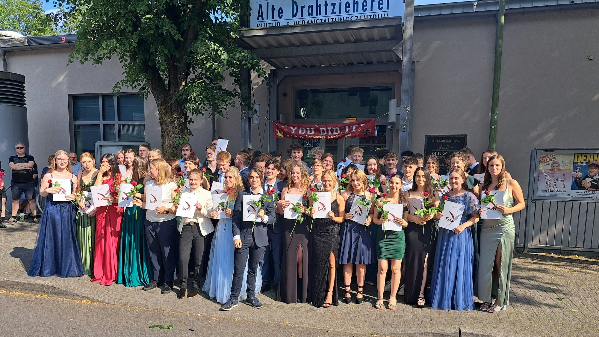 Die Schüler und Schülerinnen stehen vor der Alten Drahtzieherei und lächeln, mit Zeugnis und Rose in der Hand in die Kamera.