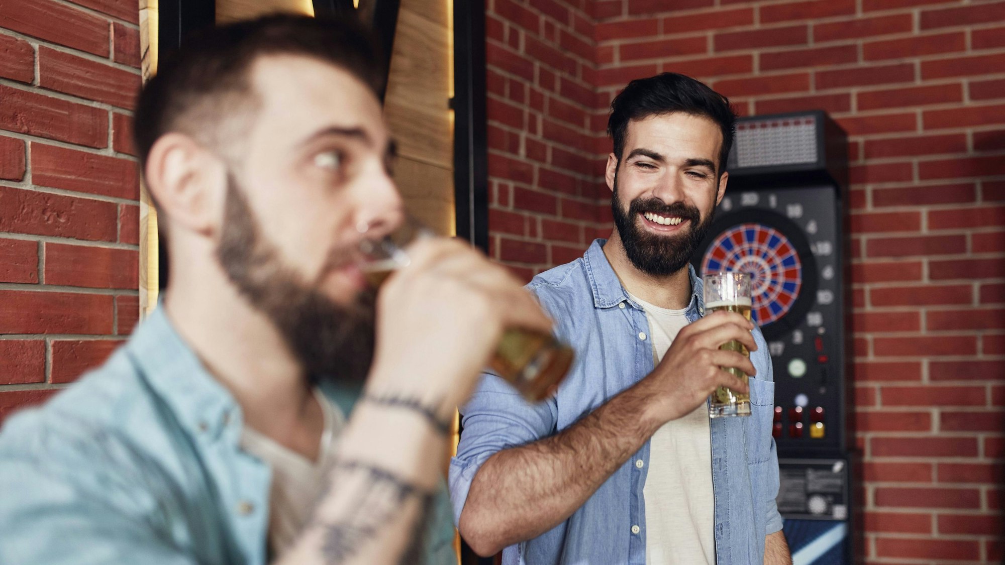 Zwei junge Männer mit dunklen Bärten stehen vor einer roten Backsteinwand und halten Biergläser in der Hand. Einer der Männer trinkt, der andere lächelt.