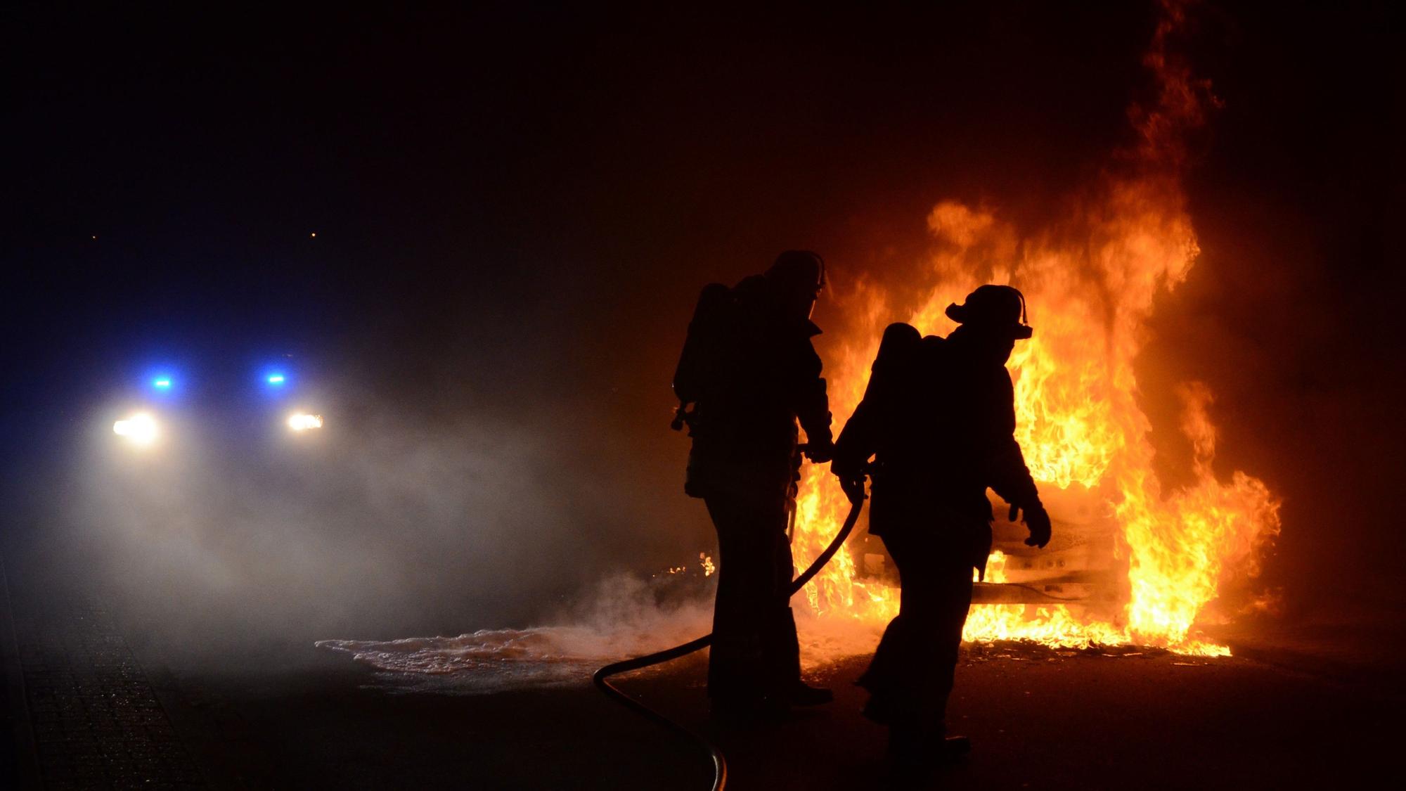 Dunkle Umrisse von zwei Feuerwehrleuten. Im Hintergrund sieht man ein brennendes Autowrack. (Archivbild)