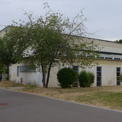 Das Bild zeigt die von Bäumen und Sträuchern umgebene Sporthalle.